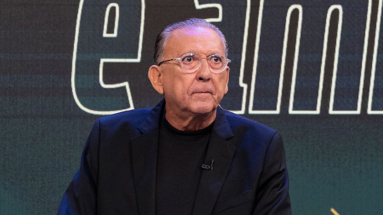 Galvão Bueno usa óculos transparentes e blazer preto em frente a painel com letras grandes iluminadas