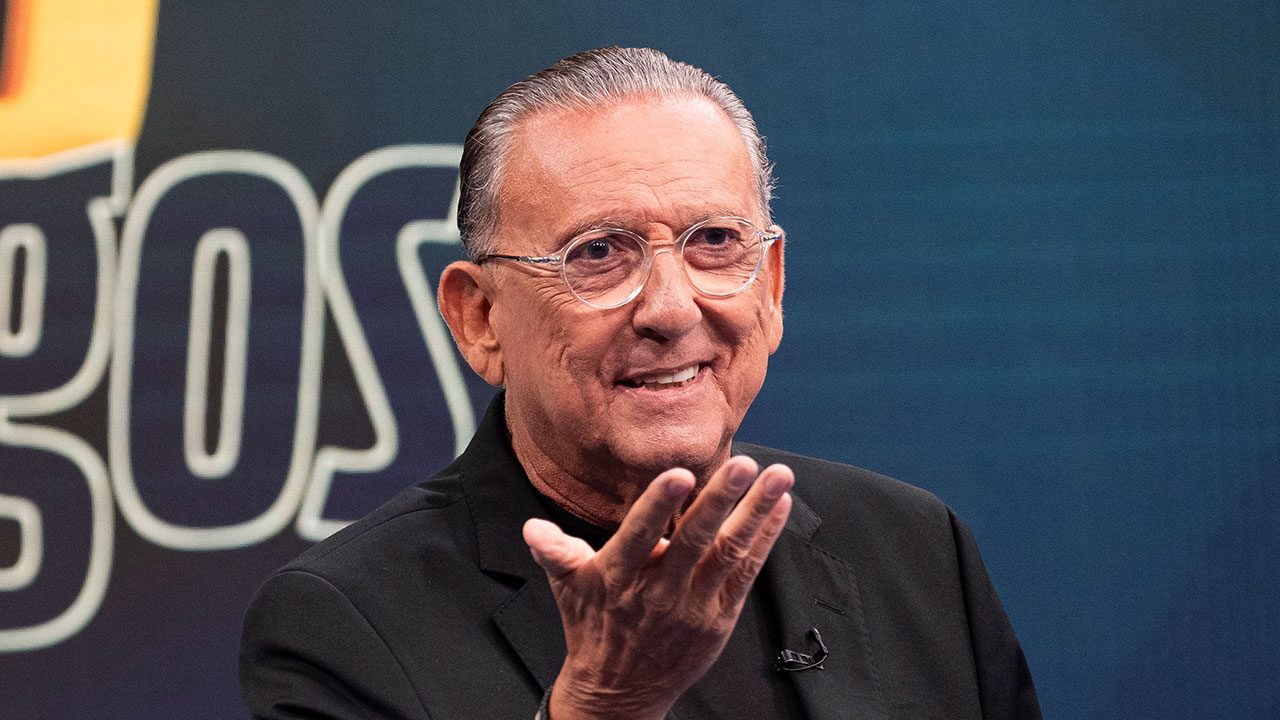 Galvão Bueno com blazer preto e óculos redondos gesticulando com a mão aberta em fundo azul de estúdio