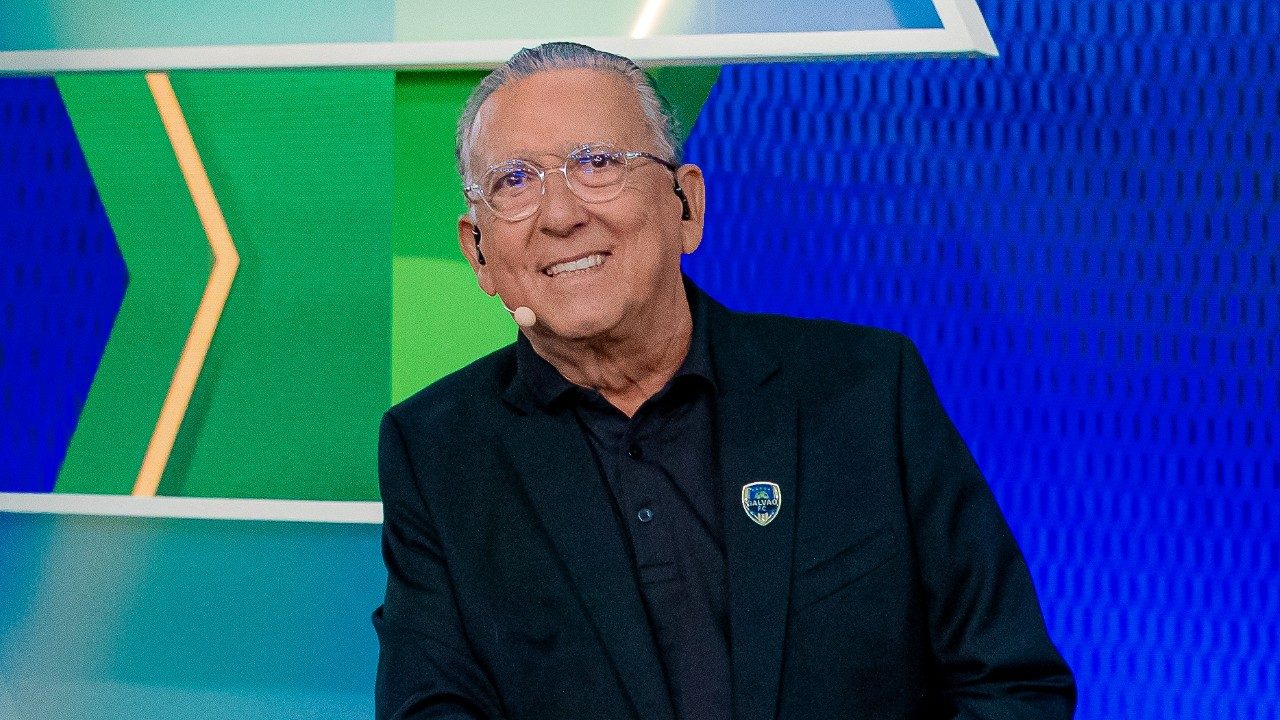 Galvão Bueno sorri usando terno escuro e microfone de ouvido em estúdio de programa esportivo na TV