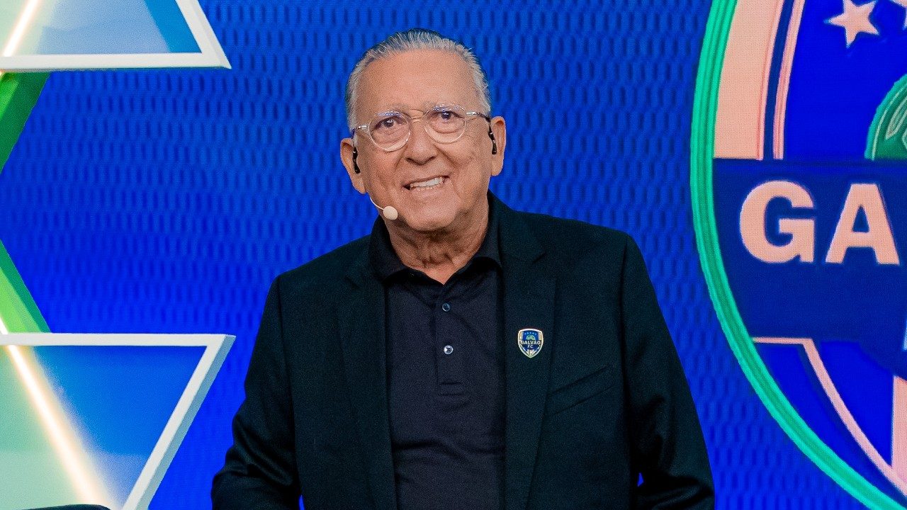 Galvão Bueno no estúdio do Galvão F.C. no SBT usando blazer escuro e microfone de lapela diante de painel azul do programa