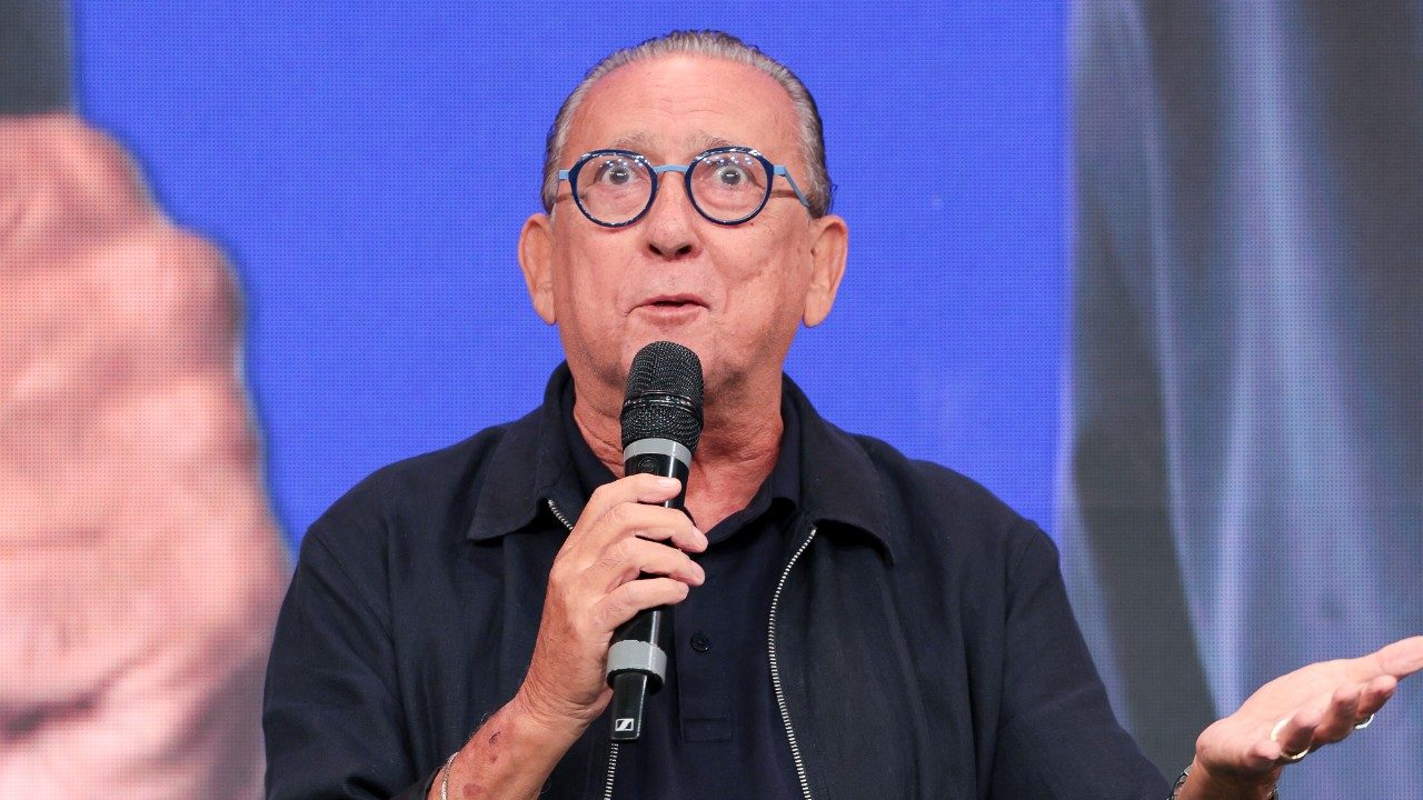 Galvão Bueno de óculos redondos segura microfone e gesticula em palco durante evento