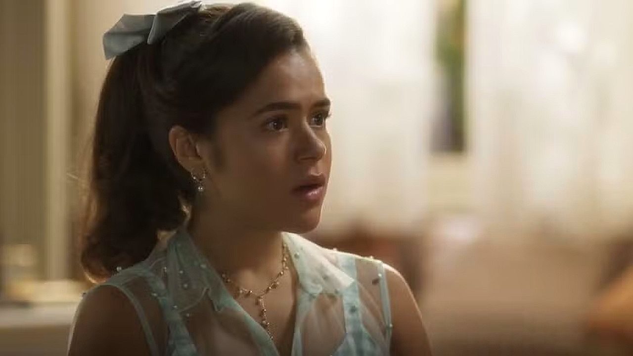 Jovem com laço no cabelo reage surpresa durante conversa em cena de novela ambientada em casa