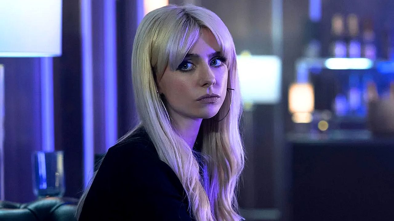 Mulher loira com expressão séria em ambiente com luz azul em cena de série ambientada em bar ou lounge