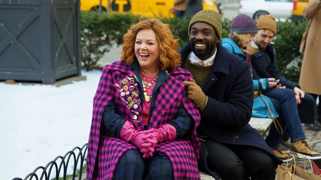 Mulher ruiva de casaco xadrez rosa sorri sentada em banco enquanto homem de gorro e casaco escuro se aproxima sorridente em praça com neve e pessoas ao fundo