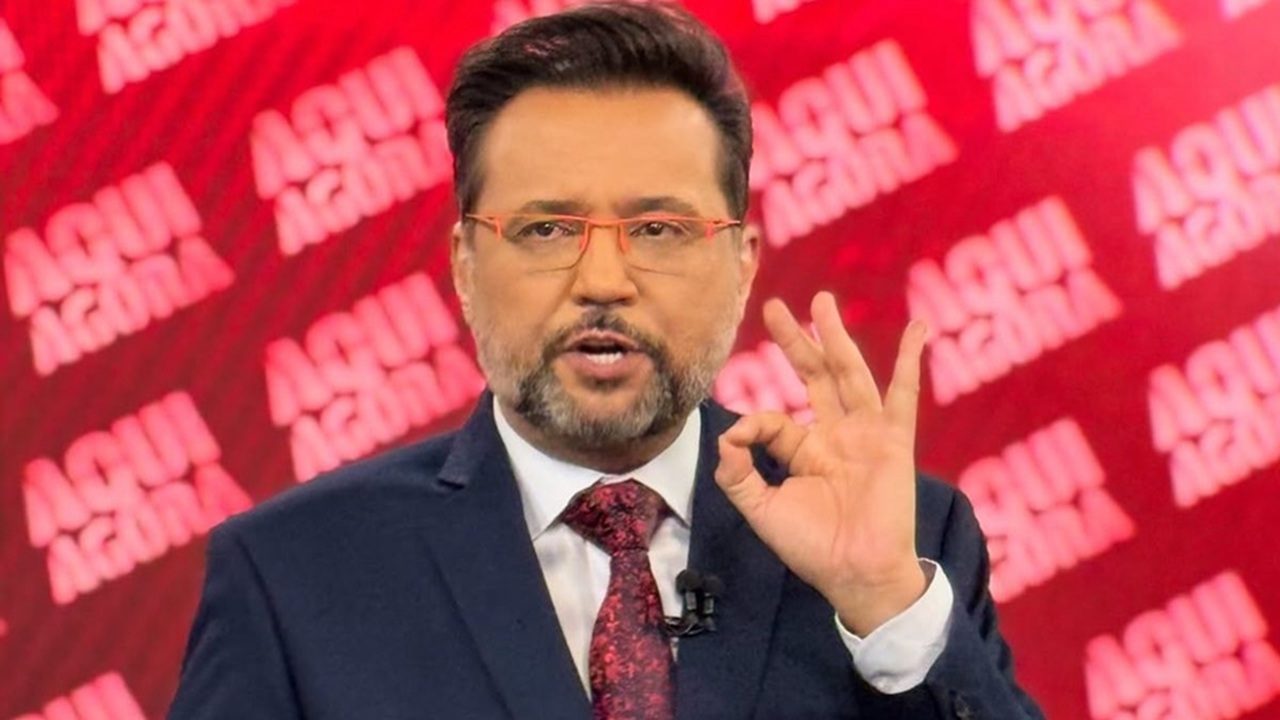 A imagem mostra Geraldo Luís, um apresentador de telejornal em estúdio, com o logotipo Aqui Agora repetido em vermelho ao fundo. Ele veste terno escuro, gravata vermelha e usa óculos de armação alaranjada. O homem faz um gesto com a mão enquanto fala, em um cenário moderno e iluminado