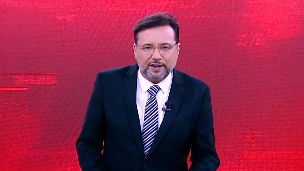 Geraldo Luís de terno escuro e gravata listrada apresenta o Aqui Agora diante do telão vermelho no SBT