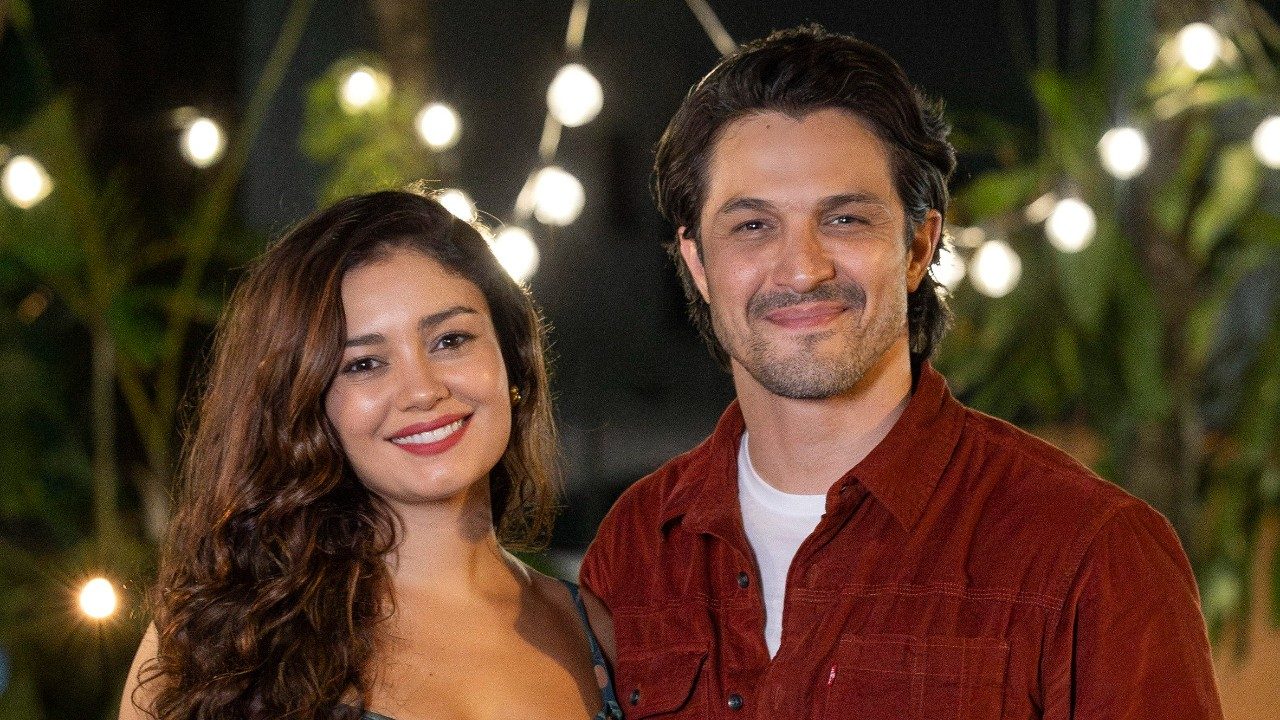 Gerluce (Sophie Charlotte) e Paulinho (Romulo Estrela) sorrindo juntos em cena da novela Três Graças com luzes ao fundo