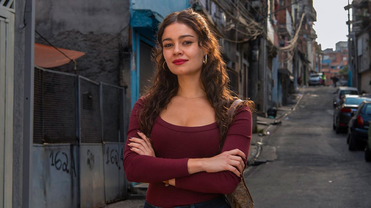 Sophie Charlotte como Gerluce com maquiagem forte e batom vermelho posa com os braços cruzados na rua de uma comunidade