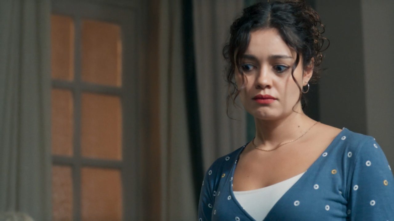 Gerluce, com expressão de preocupação está em ambiente interno com cortinas e janela ao fundo, vestindo blusa azul com pequenos círculos brancos e amarelos sobre regata branca em cena da novela Três Graças; personagem é vivida pela atriz Sophie Charlotte