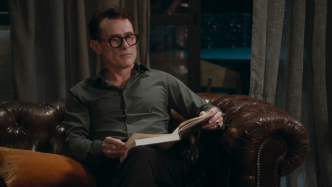 Cena da novela Volta por Cima, da TV Globo, com o personagem Gerson, interpretado por Leonardo Vieira. Ele aparece sentado em um sofá de couro marrom, segurando um livro aberto no colo, com expressão atenta.