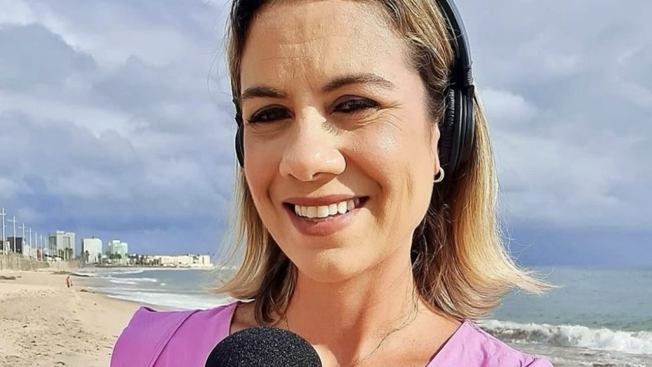 Repórter em uma praia, sorrindo para a câmera. Ela usa fones de ouvido e segura um microfone. O mar aparece ao fundo, junto com prédios ao longe sob um céu nublado.