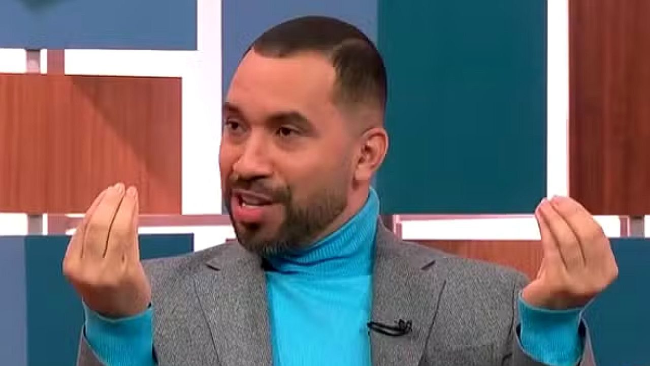 Comentarista fala gesticulando durante programa de TV em estúdio com blazer cinza e gola azul