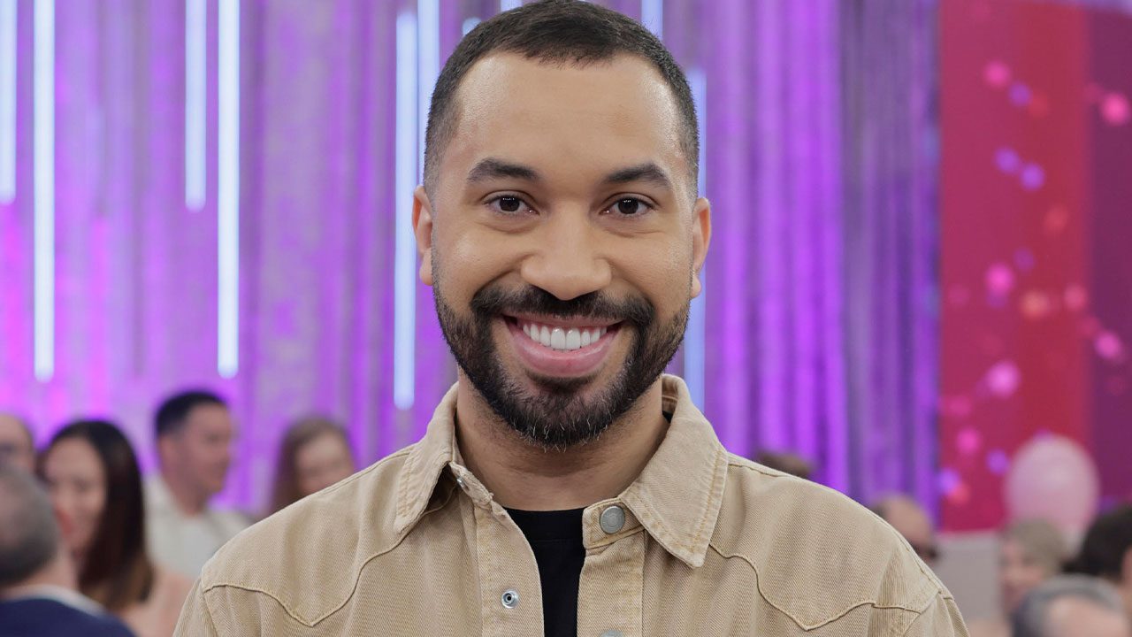 Gil do Vigor sorrindo em estúdio com iluminação roxa e plateia ao fundo