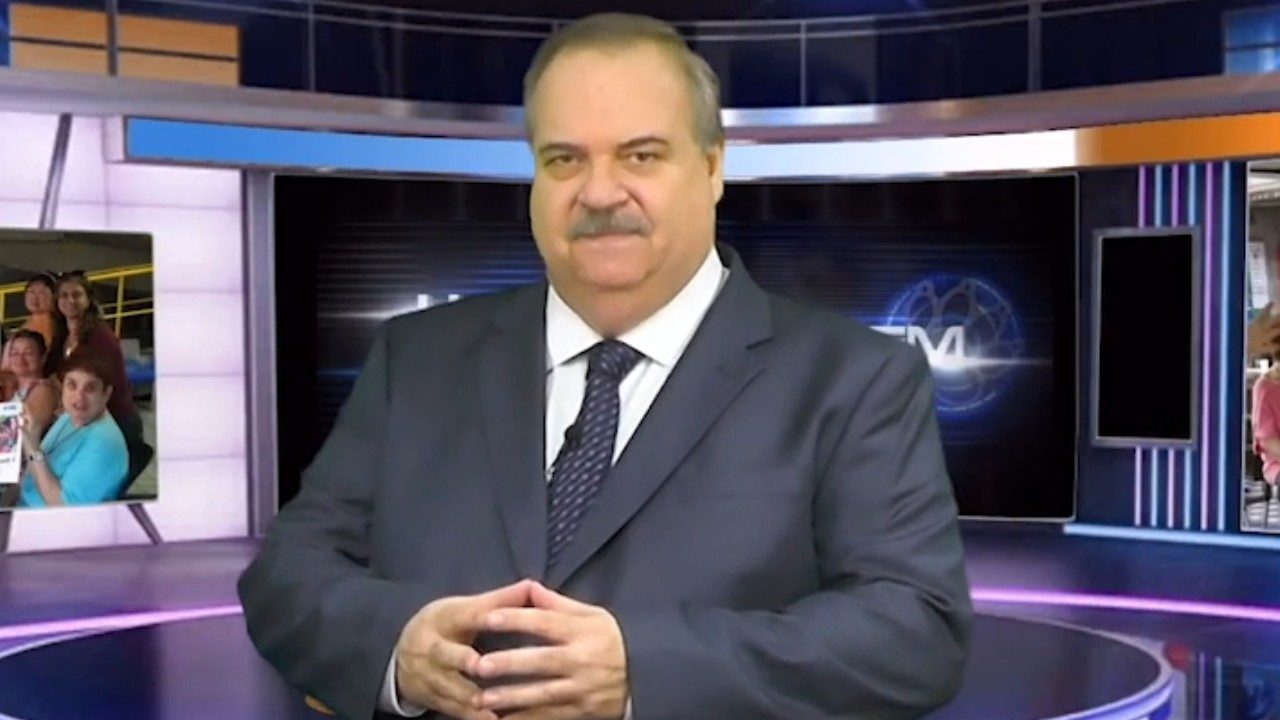 Gilberto Barros de terno azul-marinho com mãos entrelaçadas em estúdio de TV diante de telão e painéis laterais