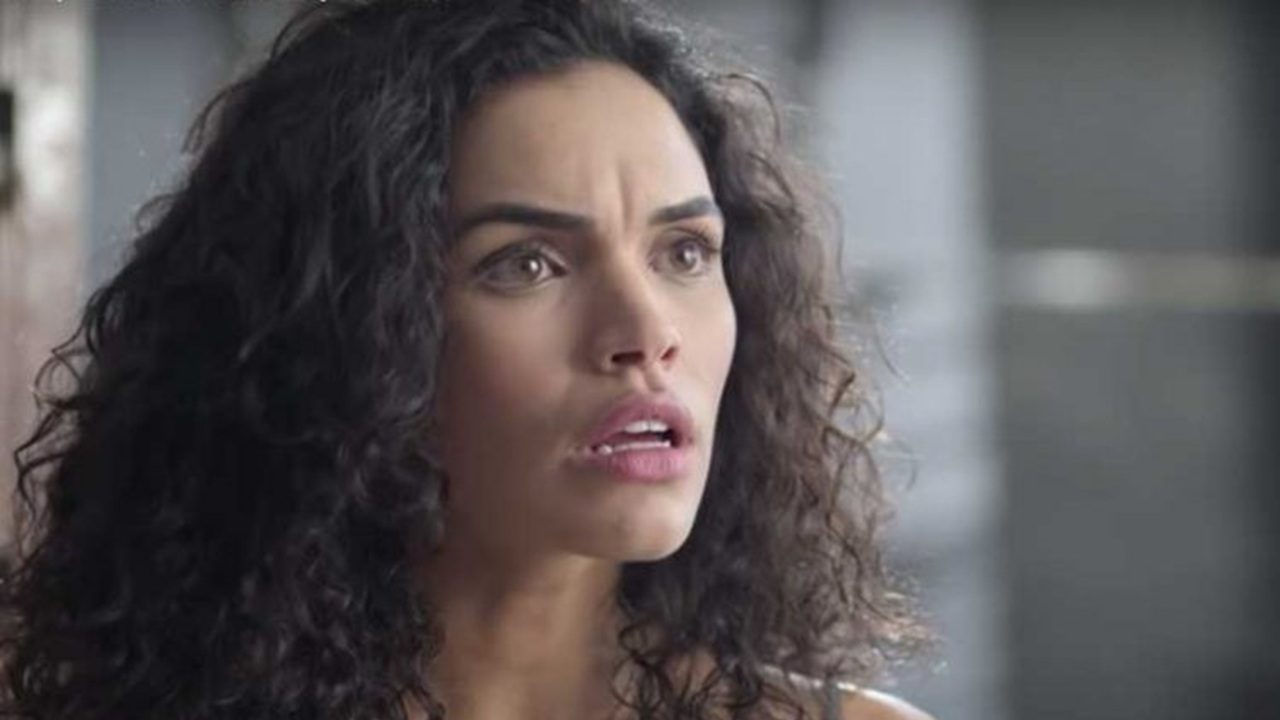 Mulher de cabelos cacheados escuros aparece em close, com expressão de surpresa e preocupação; seus olhos estão arregalados e a boca entreaberta, enquanto o fundo desfocado sugere um ambiente interno iluminado suavemente