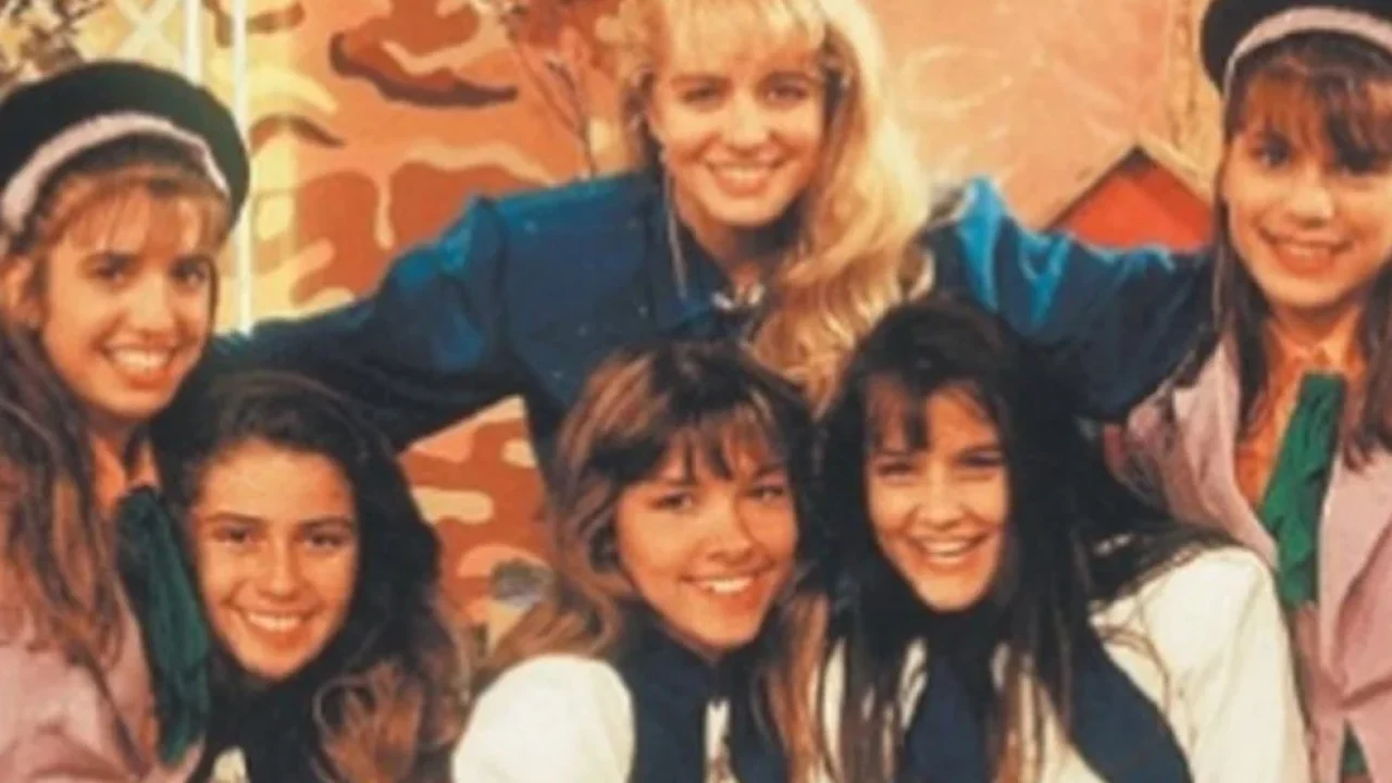 A foto parece antiga (provavelmente anos 1980 ou início dos 1990), com seis jovens e uma mulher adulta ao centro, todos sorrindo e muito próximos, passando uma sensação de grupo unido. Eles usam roupas parecidas, com lenços no pescoço e alguns chapéus, o que sugere um elenco infantil/juvenil, possivelmente de um programa de TV, grupo musical ou projeto artístico da época. A mulher ao centro parece ter uma posição de liderança ou destaque, com os braços abertos sobre os jovens.