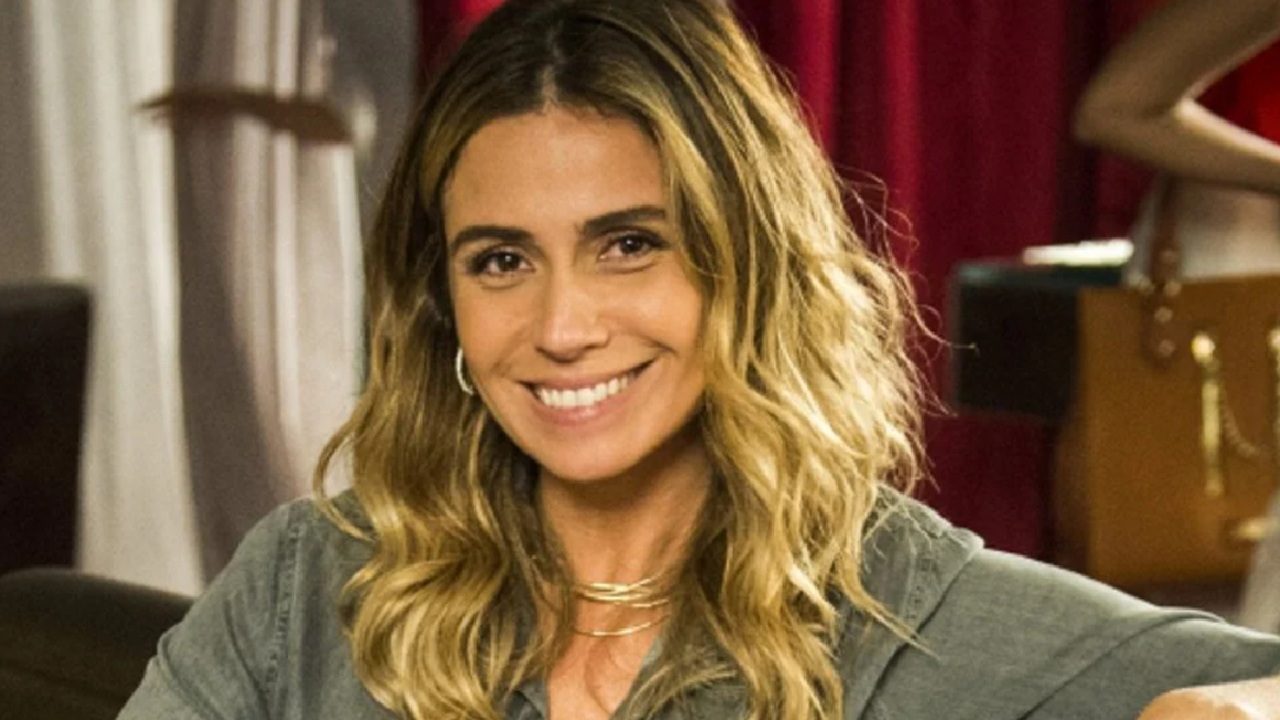 Mulher de cabelos loiros ondulados e blusa cinza sorri em ambiente interno com cortinas vermelhas ao fundo