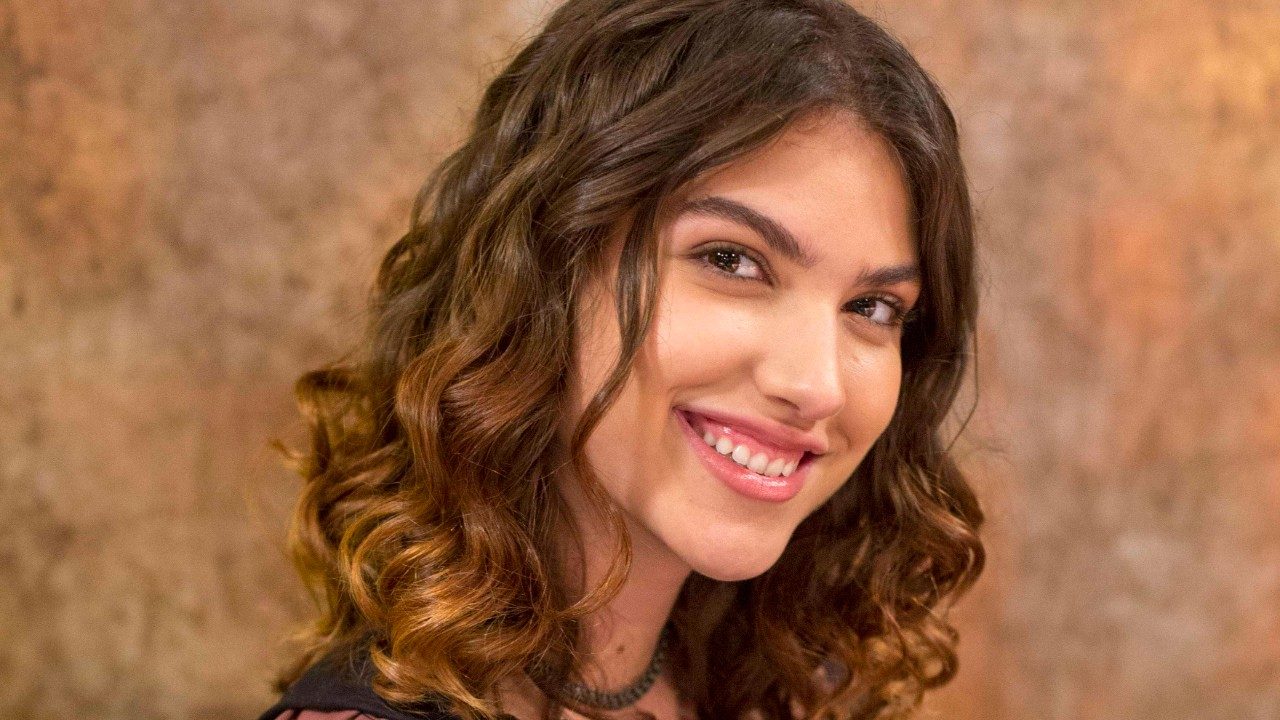Giovanna Grigio sorri em retrato com cabelo ondulado e fundo neutro em tons terrosos