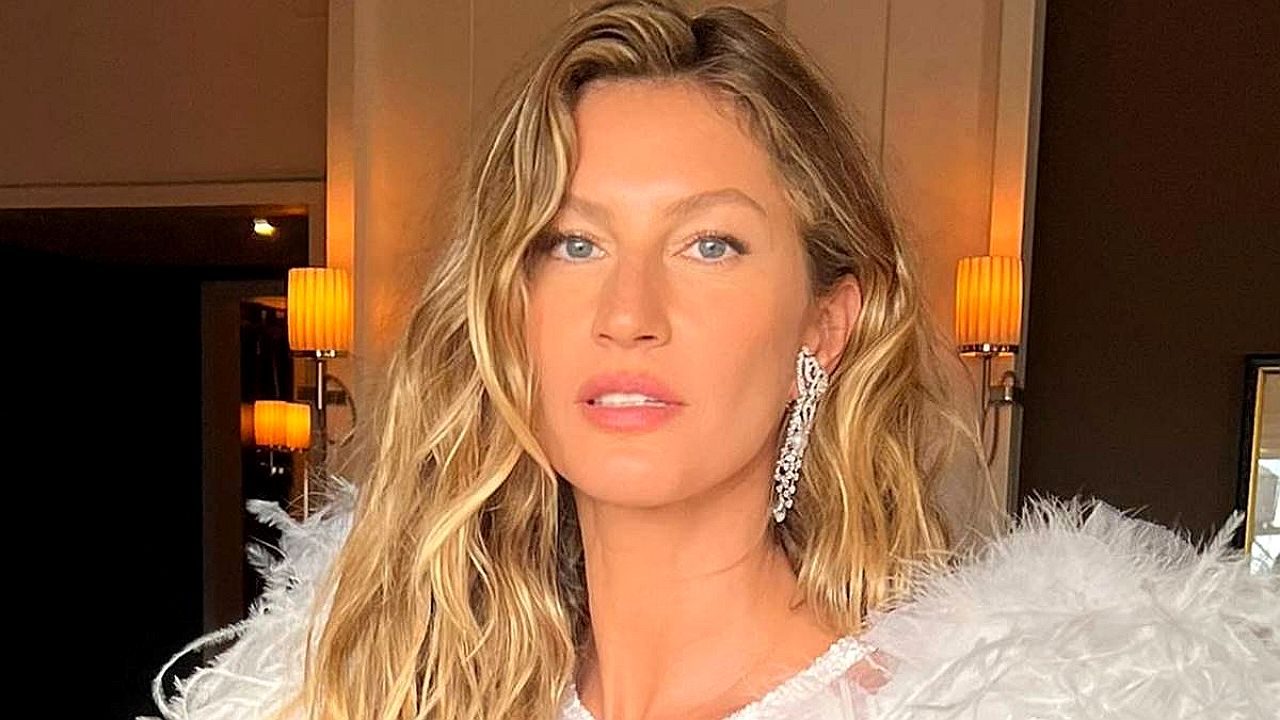 Gisele Bündchen com vestido claro e detalhe de plumas posa em ambiente interno com iluminação quente