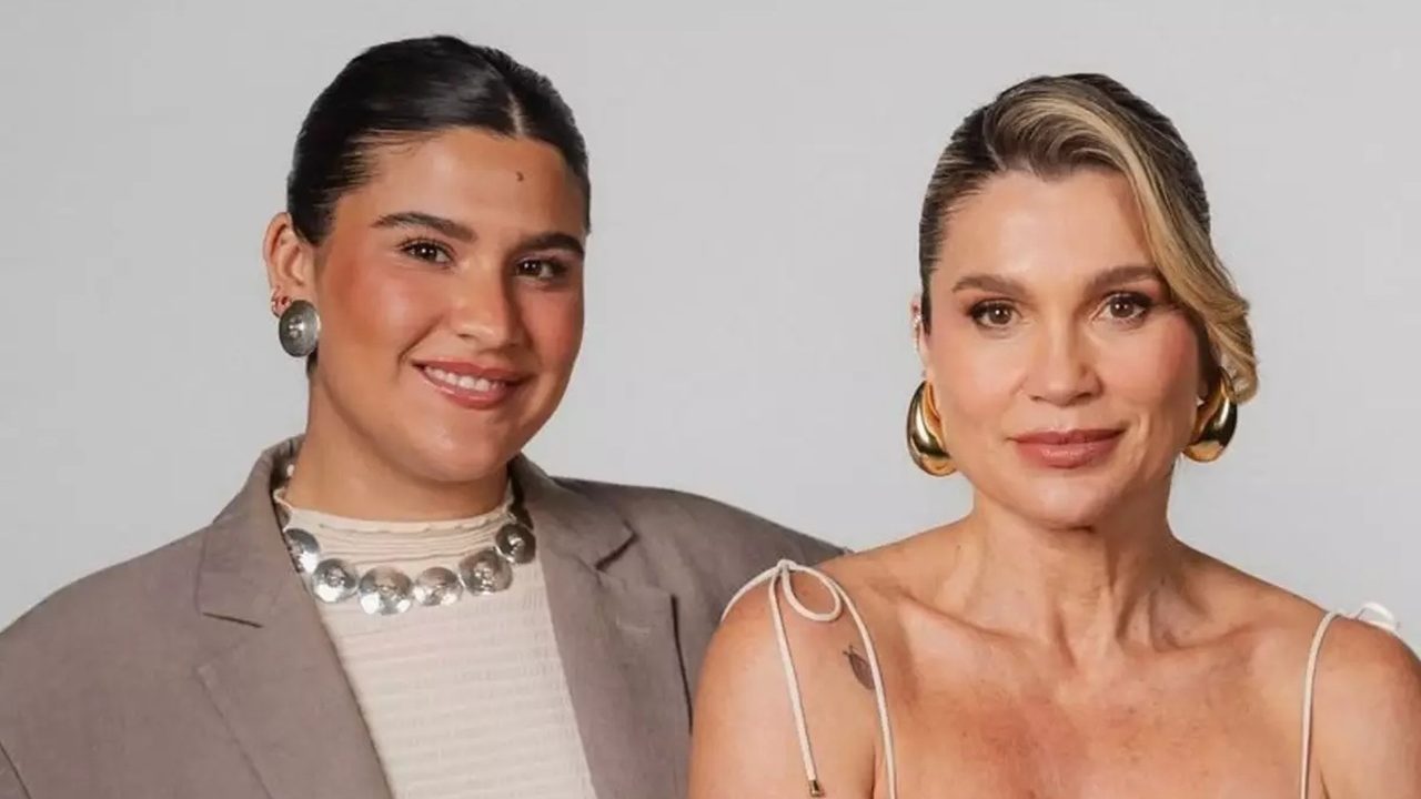 Duas mulheres posam lado a lado diante de fundo neutro. A mulher à esquerda veste blazer bege, blusa clara e colar metálico, sorrindo suavemente. A mulher à direita usa vestido de alças finas e brincos dourados grandes, olhando para a câmera com expressão serena