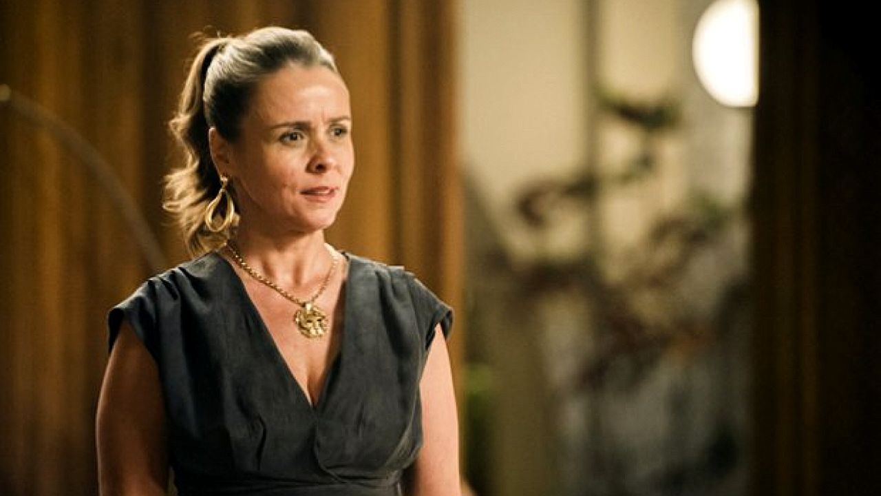 Mulher de vestido preto e rabo de cavalo fala com expressão séria em cena de novela em ambiente interno elegante