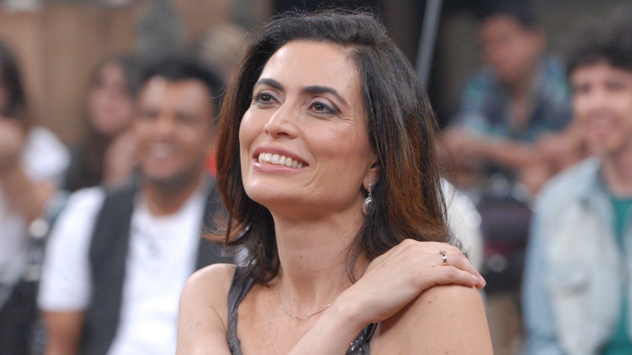 Giuliana Morrone sorri em estúdio de TV com plateia desfocada ao fundo e mão apoiada no ombro