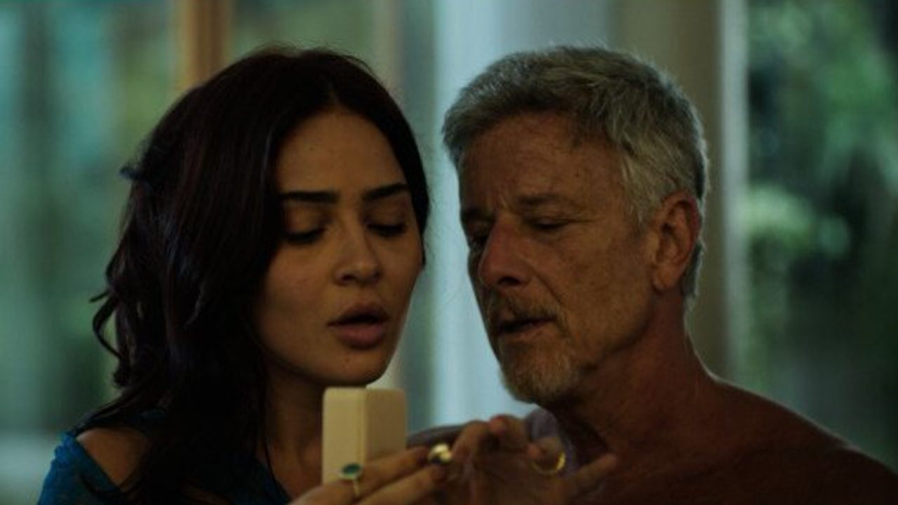 A imagem mostra duas pessoas em um ambiente interno. A mulher, de cabelos escuros e soltos, está segurando uma caixinha aberta — possivelmente de joias ou anel — enquanto o homem, de cabelos grisalhos e barba, observa atentamente o objeto. A cena parece íntima e carregada de emoção