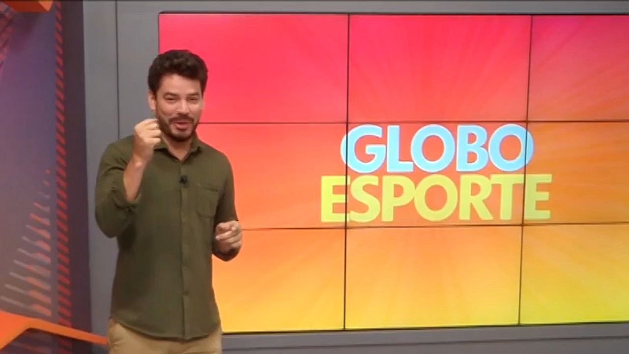 Apresentador do Globo Esporte gesticula no estúdio diante de telão com logotipo do programa