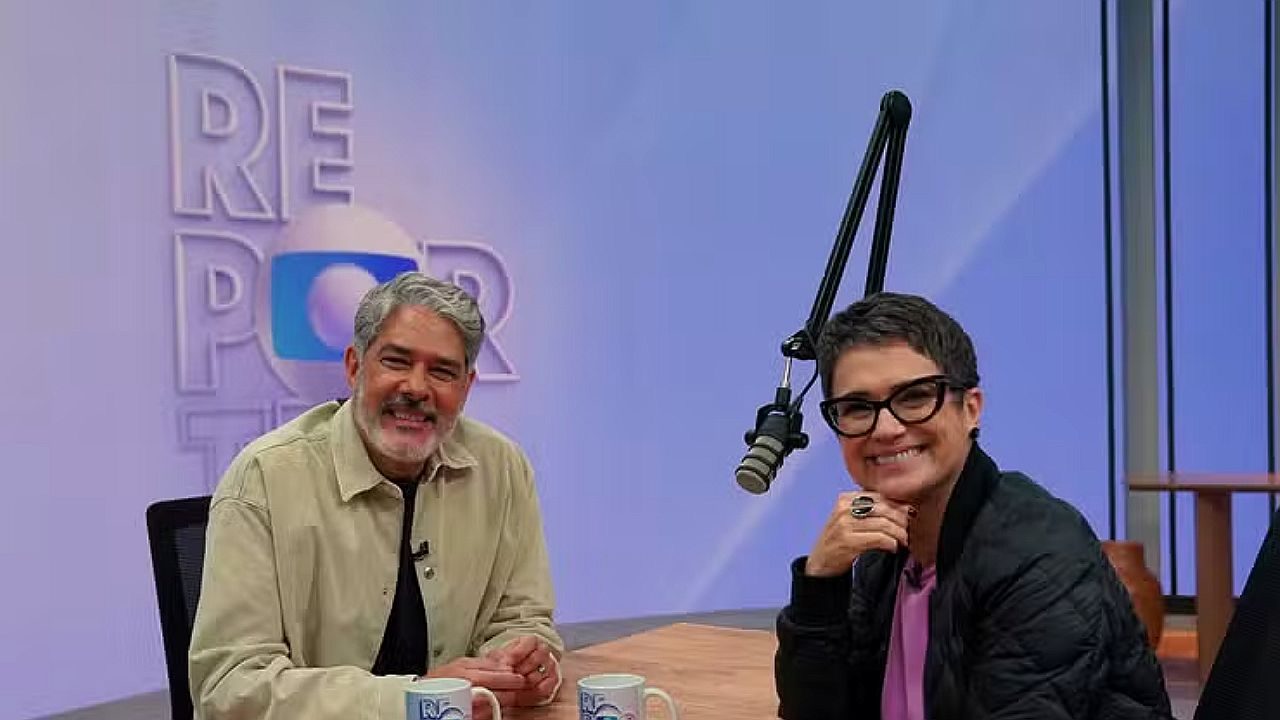 William Bonner e Sandra Annenberg posam sorrindo em estúdio de podcast com microfone suspenso e canecas sobre a mesa