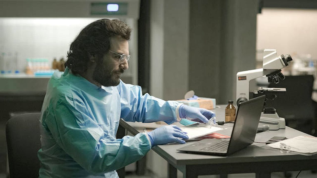 Homem com roupa de proteção analisa amostras em laboratório com microscópio e notebook sobre a mesa