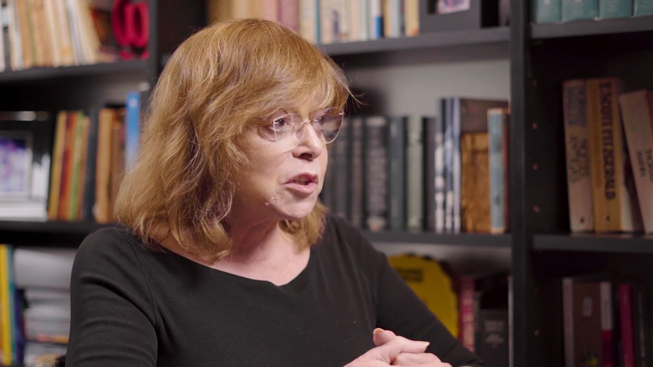 Gloria Perez sentada e falando em ambiente com estante de livros ao fundo