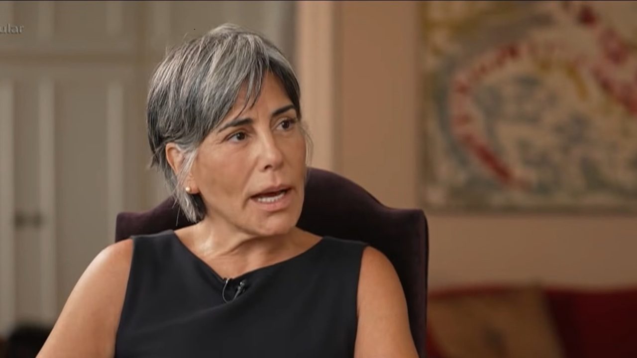 Mulher de cabelo curto grisalho participa de uma entrevista em ambiente interno, usando blusa preta sem mangas e falando enquanto olha para o lado; ao fundo, aparecem móveis e um quadro colorido desfocado.