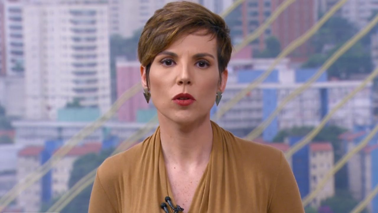 Glória Vanique com expressão séria durante apresentação de telejornal com fundo de cidade ao amanhecer