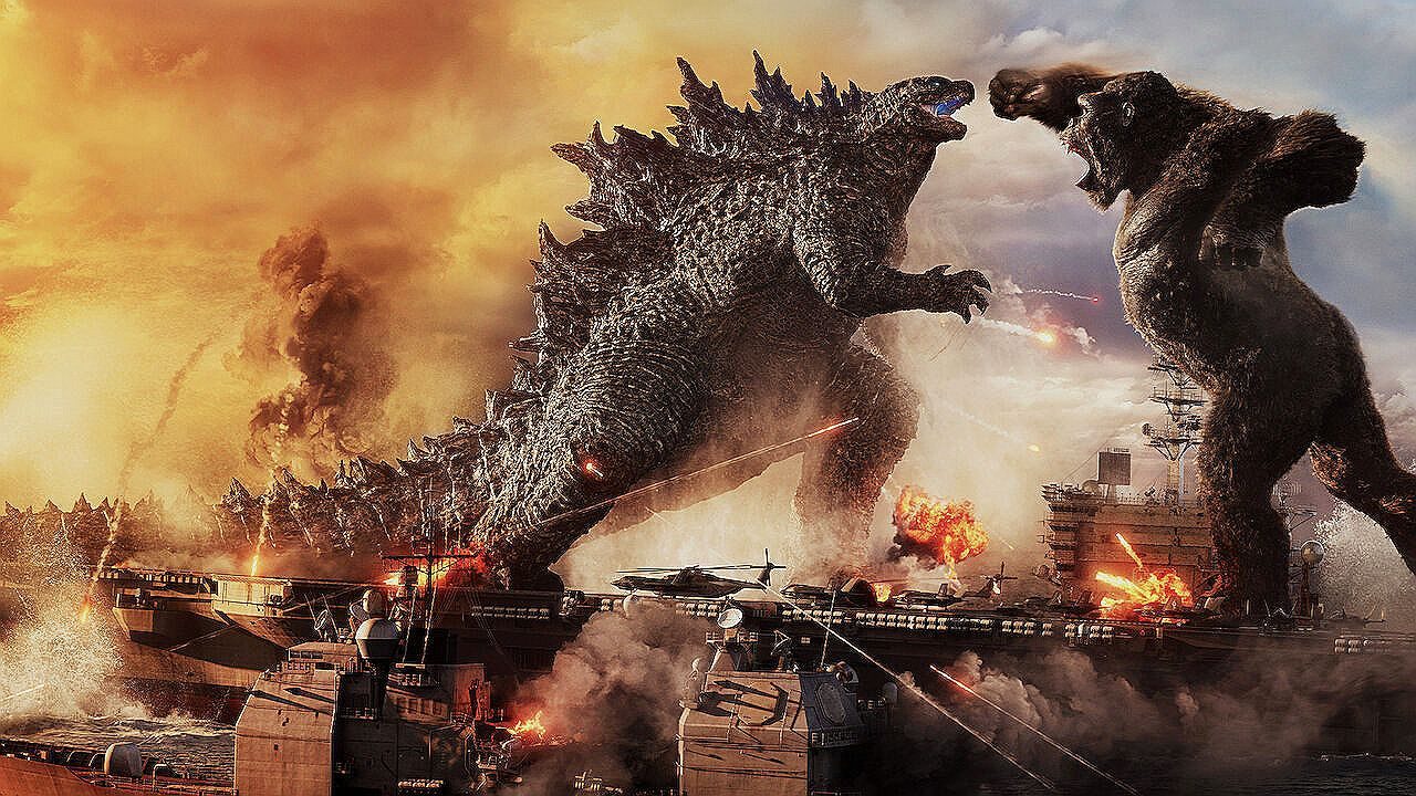 Cena épica mostra dois monstros gigantes em batalha sobre o mar, próximos a navios militares e porta-aviões. À esquerda, uma criatura reptiliana enorme com placas dorsais afiadas avança rugindo. À direita, um gigantesco gorila também ruge e ergue o braço para atacar. Explosões, fumaça e helicópteros ao redor intensificam o clima de destruição, enquanto o céu alaranjado sugere pôr do sol ou cenário apocalíptico.