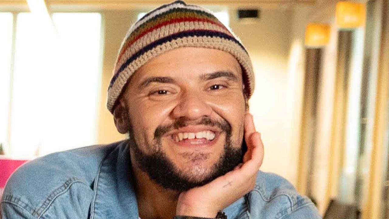 Gominho sorri para a câmera usando um gorro de crochê listrado e jaqueta jeans, apoiando o queixo na mão esquerda