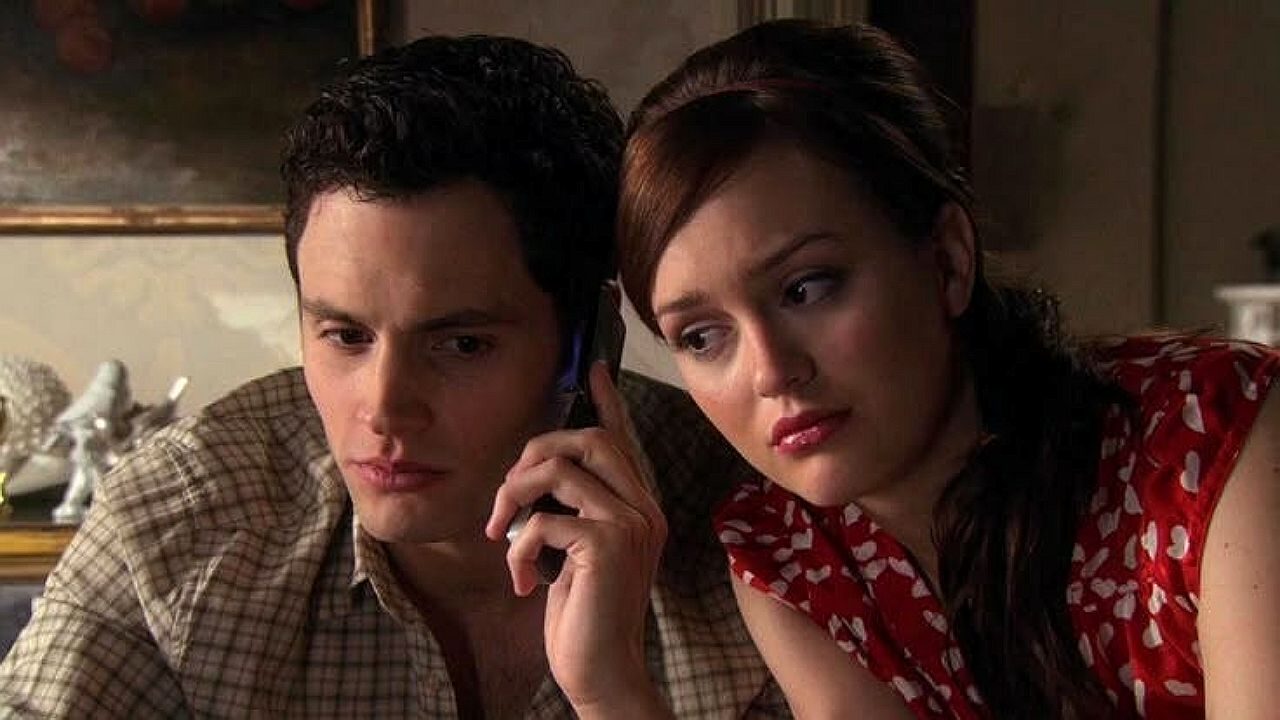 Um homem segura um celular junto ao ouvido com expressão séria, enquanto uma mulher apoia a cabeça em seu ombro e observa com olhar preocupado. Eles estão em ambiente interno aconchegante, com decoração ao fundo, sugerindo cena dramática de série ou filme.