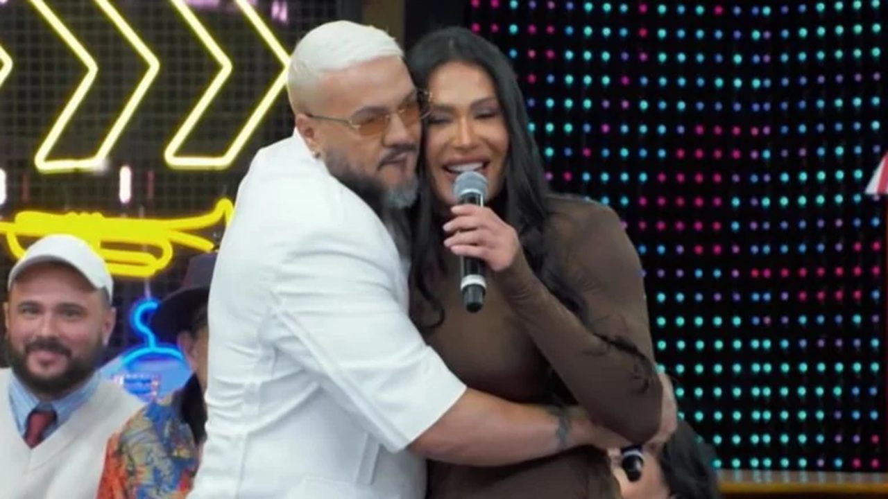 A imagem mostra um homem e uma mulher se abraçando em um palco colorido. O homem, de cabelo e barba grisalhos e óculos alaranjados, veste roupa branca. A mulher segura um microfone e usa um vestido marrom, sorrindo enquanto canta ou fala.