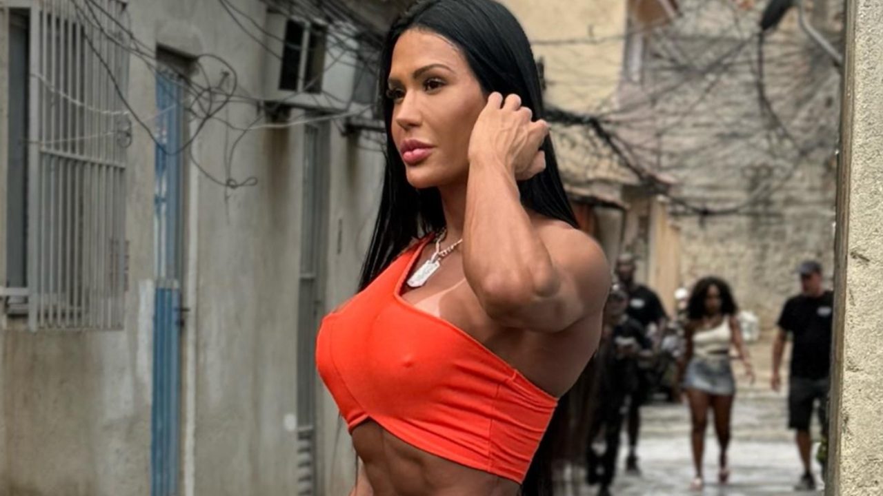 Mulher de cabelos longos e pretos usa top laranja de um ombro só e colar prateado enquanto caminha por viela com paredes de concreto e fios expostos, com outras pessoas ao fundo