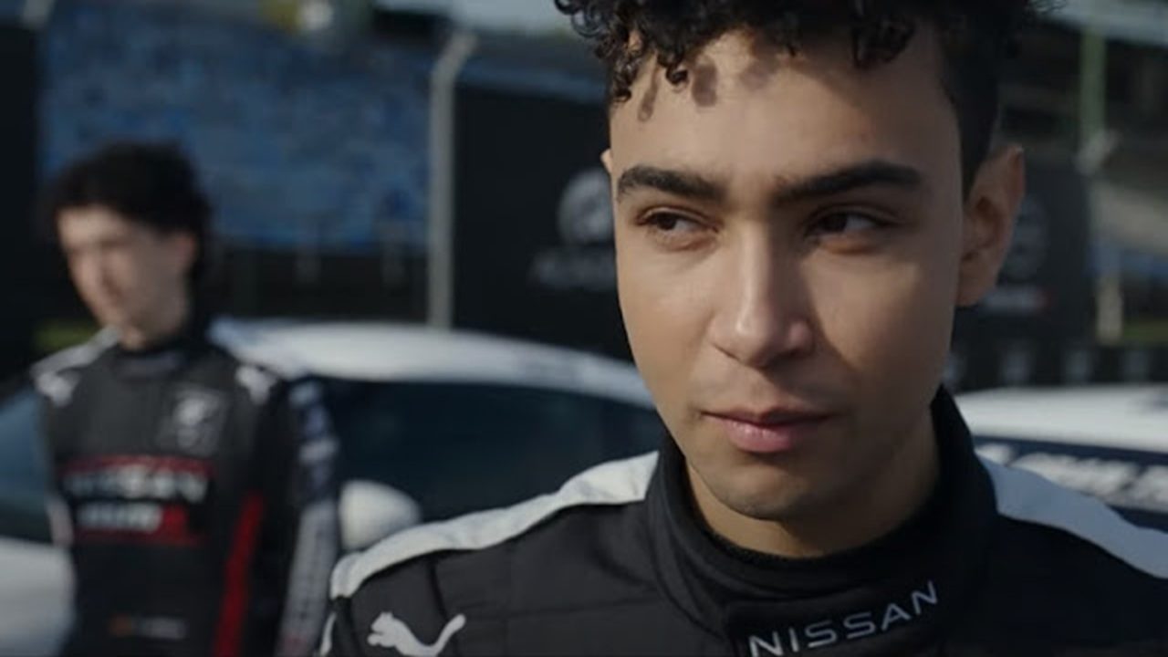 Homem jovem com expressão confiante aparece em primeiro plano vestindo um macacão de piloto preto com detalhes brancos e o logotipo da Nissan. Ao fundo, outro piloto com traje semelhante está desfocado, em frente a carros de corrida brancos estacionados em um autódromo sob luz natural.