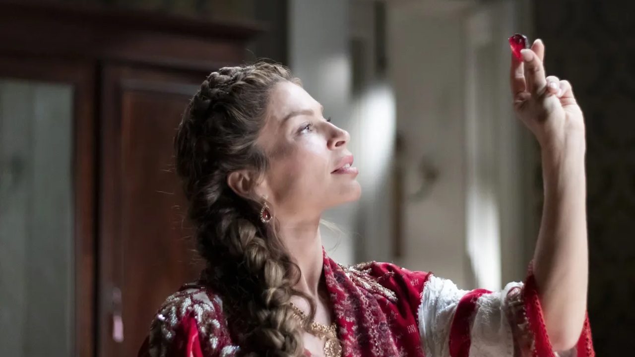 Mulher com vestido vermelho de estilo clássico e cabelo trançado observa atentamente uma pequena joia vermelha que segura entre os dedos, em ambiente interno com móveis de madeira ao fundo.