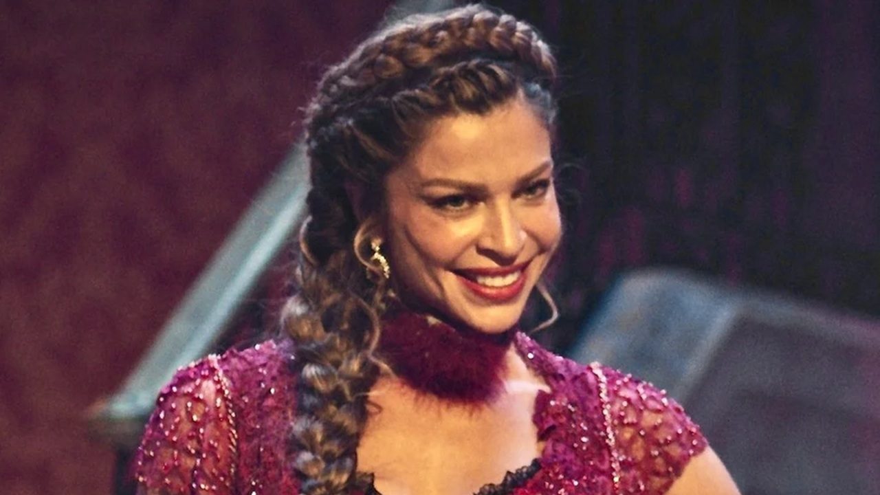 Mulher com vestido vermelho bordado e trança no cabelo sorri em ambiente com iluminação teatral