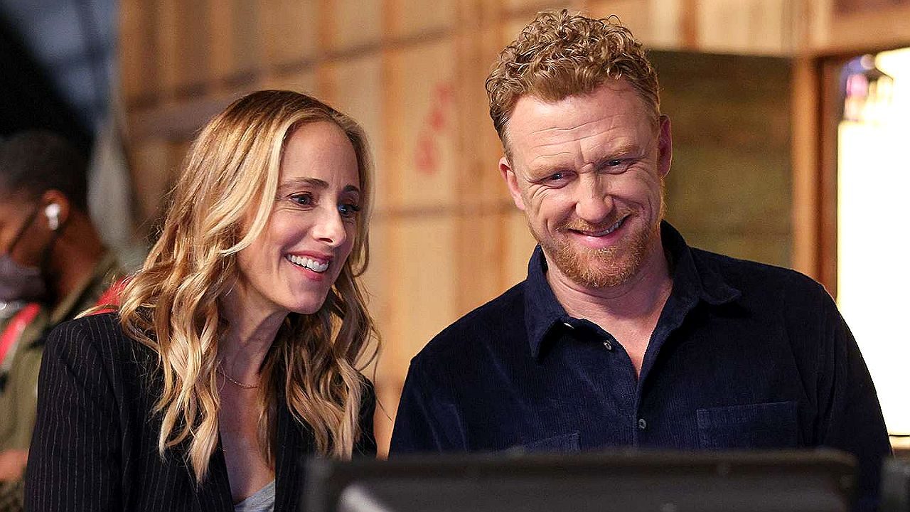 Homem e mulher sorriem enquanto observam tela em ambiente de trabalho em cena de série médica Grey’s Anatomy