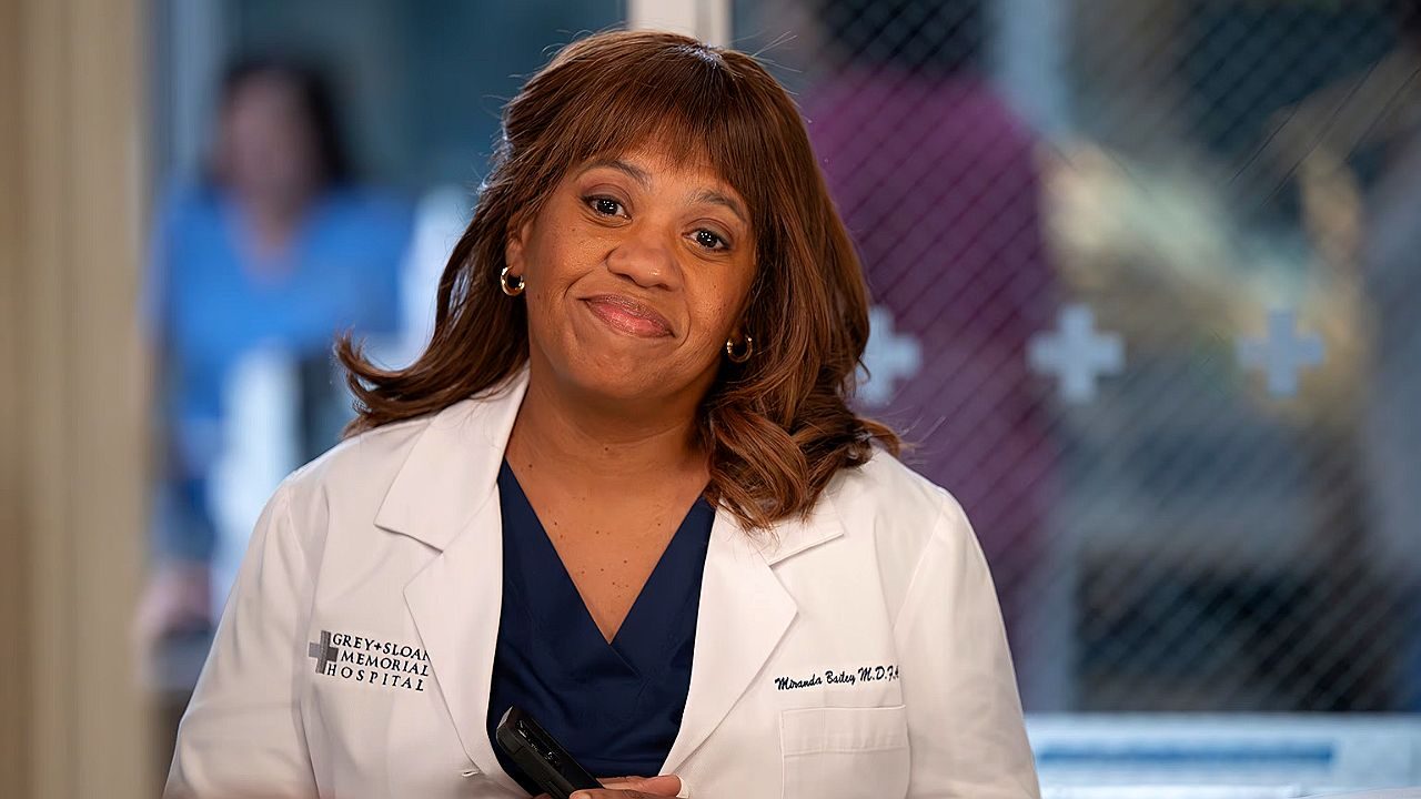 Mulher de jaleco médico sorri em hospital com identificação Grey Sloan Memorial Hospital visível no uniforme