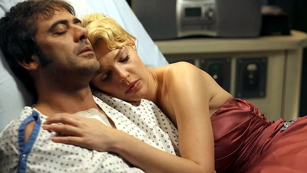 Um homem está deitado em uma cama de hospital usando roupa hospitalar, enquanto uma mulher de vestido vermelho se deita ao lado dele e apoia a cabeça em seu peito, em um momento íntimo e emocional.