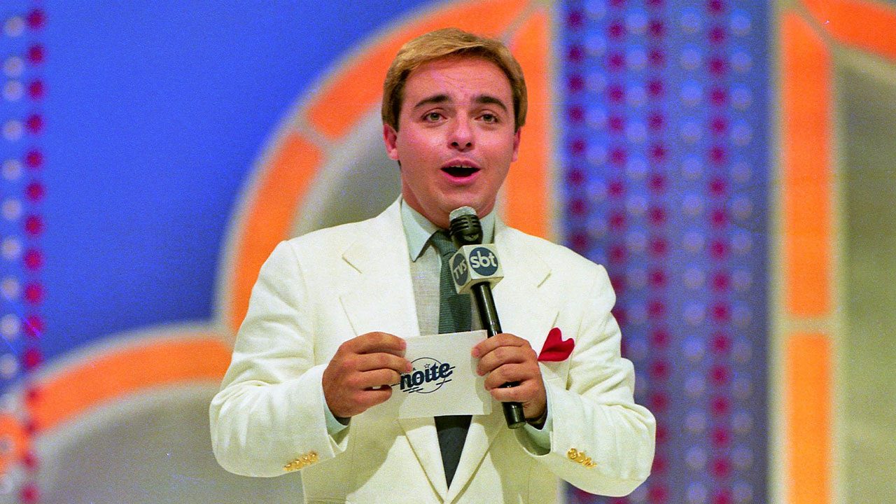 Gugu Liberato de terno branco com microfone do SBT apresenta o programa Viva a Noite segurando ficha com logo da atração