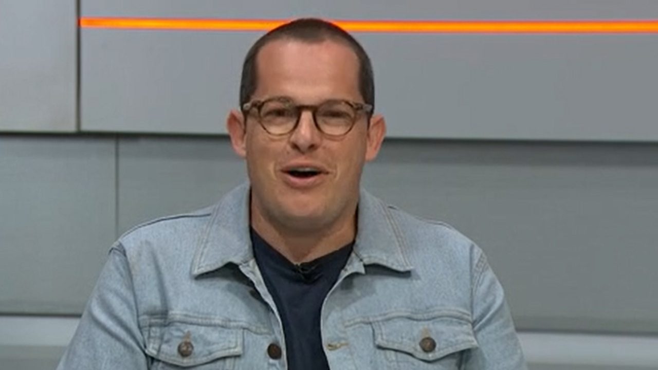 Homem de óculos e jaqueta jeans fala no estúdio de programa esportivo com painel cinza e faixa laranja ao fundo