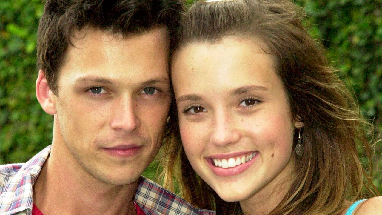 Guilherme Berenguer e Juliana Didone como Gustavo e Letícia em Malhação sorrindo juntos em área externa com vegetação ao fundo