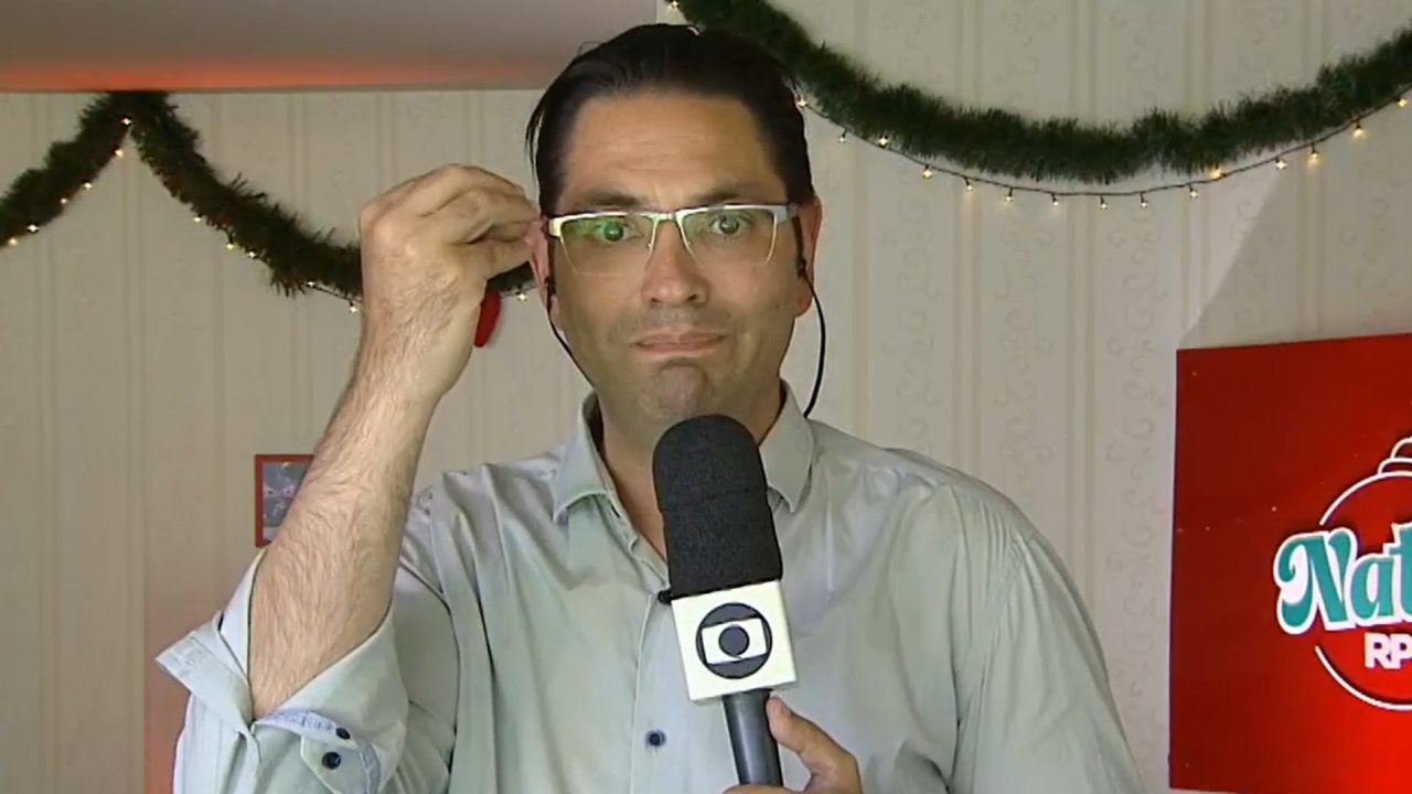 A imagem mostra Guilherme Rivaroli, segurando um microfone com o logotipo da Globo, durante uma entrada ao vivo. Ele usa óculos, uma camisa clara e está em um ambiente decorado com enfeites natalinos, mostrando que a gravação ocorreu durante o período de festas