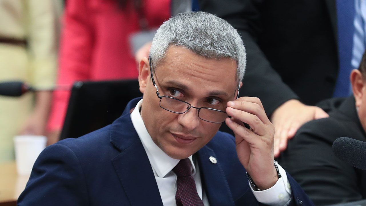 Deputado Gustavo Gayer com cabelo grisalho, vestindo terno azul marinho e ajustando os óculos em sessão da Câmara