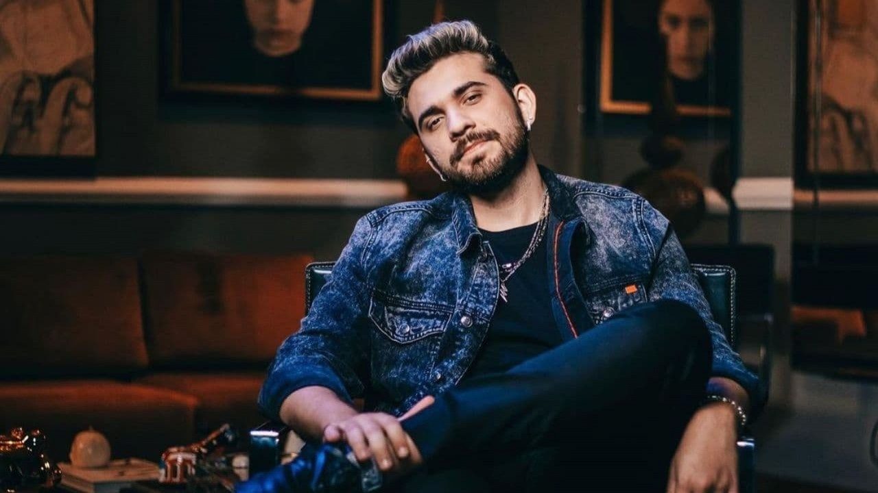 Homem jovem sentado de forma descontraída em uma poltrona, com expressão confiante e leve sorriso. Ele usa jaqueta jeans escura, camiseta preta e colar. O ambiente ao fundo é sofisticado, com sofá marrom e quadros escuros nas paredes.